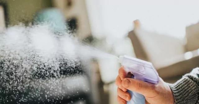 Hand spraying mist from a spray bottle with a blurred background of indoor furniture.