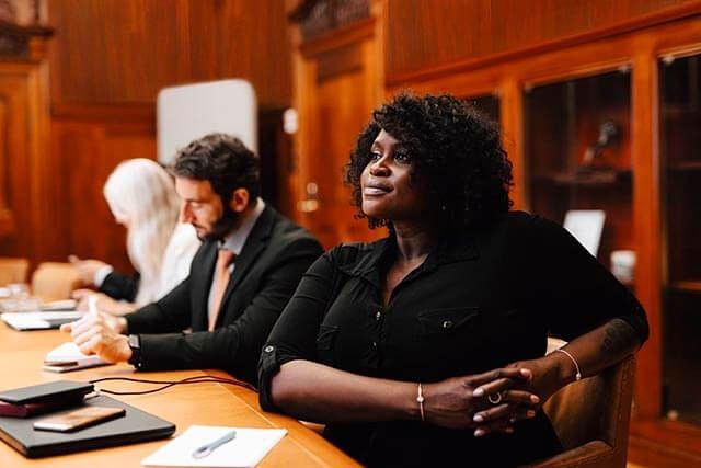 Three people sit at a wooden conference table. The woman in the foreground looks attentively forward, while a man and another woman are taking notes. The room has wooden paneling and bookshelves in the background.