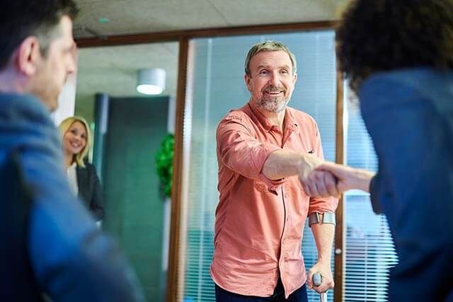 A man with a crutch in his left arm is shaking hands with someone else in an office setting.