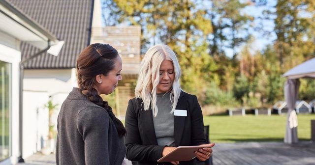Two women outside, discussing with one holding a tablet. Lawn and trees in the background.