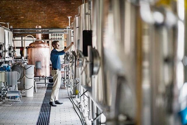 Person operating or inspecting brewing equipment in a spacious brewery with tanks and a copper vat.