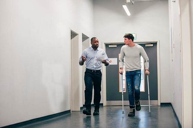 Two men, one using crutches, walk side by side down a well-lit, empty hallway.