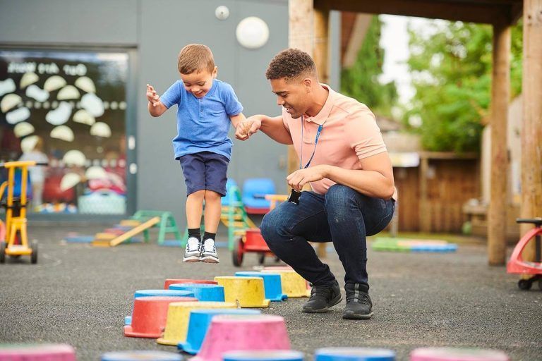 Male nursery worker holding a boy's hand as he jumps on stepping blocks in a playground.