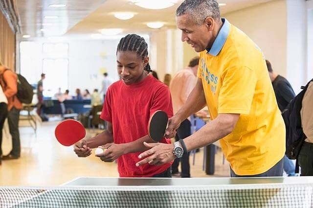 Man teaching table tennis to a young person in a well-lit sports community centre.