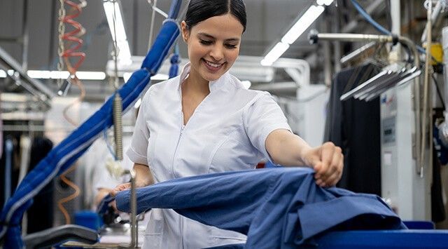 Woman wearing a white uniform working in a laundry facility, holding a shirt and operating a pressing machine.