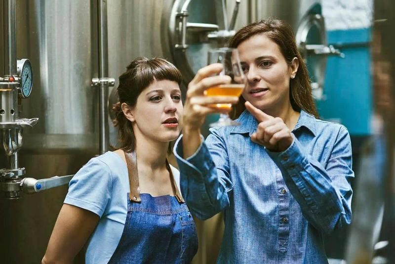 Two women examining a beer sample inside a brewery with stainless steel tanks in the background.