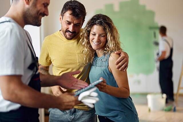 Couple with painter looking at a tablet in a room. A painter in the background is painting one of the walls green.
