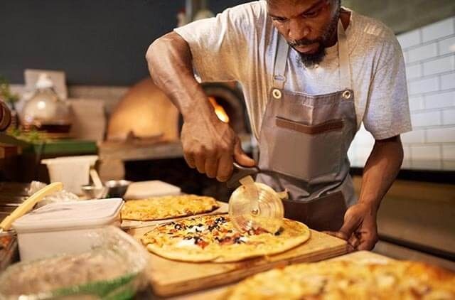 Takeway chef slicing freshly baked pizza in a kitchen with multiple pizzas on the counter.