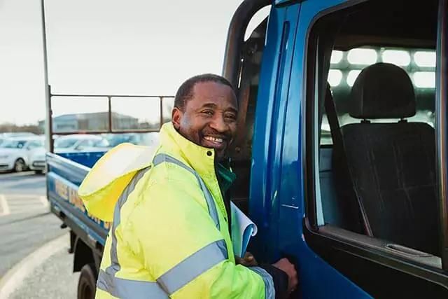 A smiling man in a hi-vis is outside of a blue truck parked in car park.