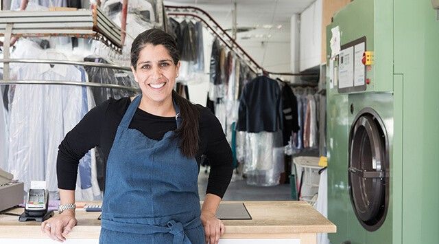 A launderetter owner wearing a blue apron smiles in front of the counter with clothing and machines in the background.