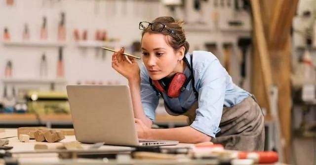 Person leaning over a laptop in a workshop, holding a pencil, surrounded by tools and equipment.