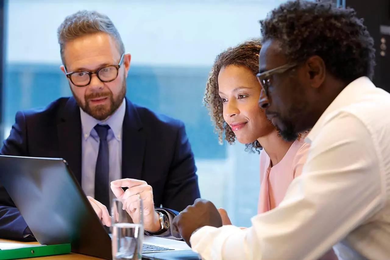 Three professionals sitting at a meeting table, discussing, with a glass of water on the table and a laptop open.