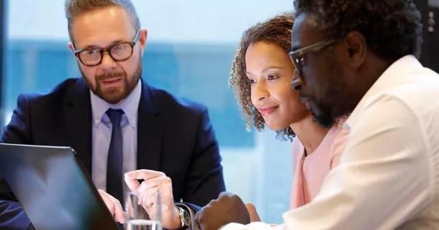 A group of three professionals speaking, all seated around a table looking at a laptop.