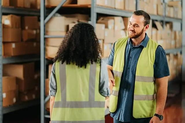 Warehouse staff in high-vis vests shaking hands in front of shelves with boxes.