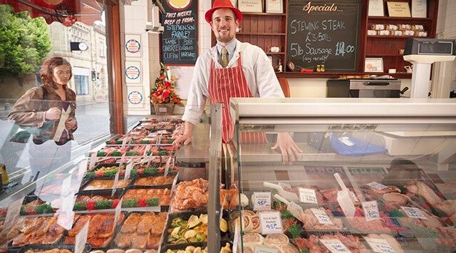 Butcher in a striped apron behind a meat display with a chalkboard listing prices in the background.