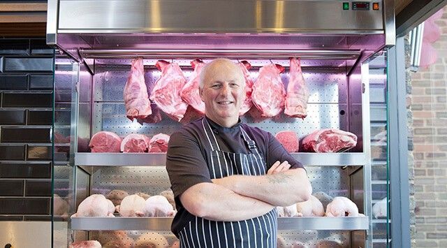 Butcher in striped apron standing in front of neatly arranged meats.
