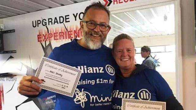 Two people wearing blue shirts with company logos hold certificates of achievement. They are smiling in an indoor setting with promotional posters on the wall behind them.