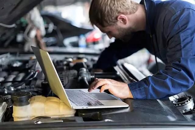 A mechanic in a blue jumpsuit is working under the hood of a car. He is using a silver laptop, which rests on the engine. His focus is on the engine, possibly diagnosing an issue or looking up repair information.