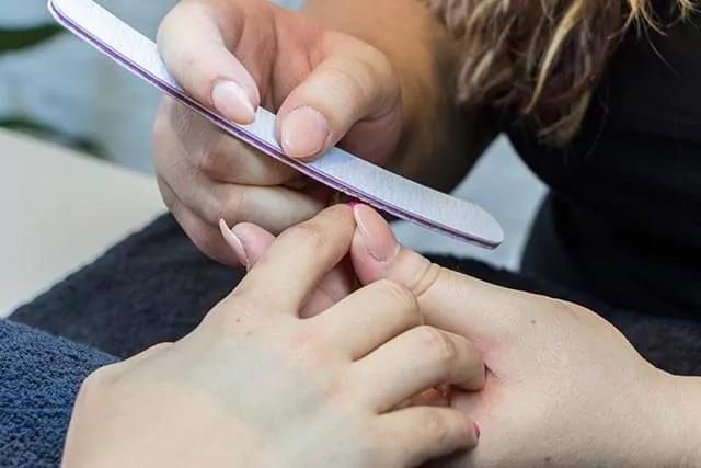 A nail tech shaping nails using a file on another's hand, resting over a dark towel.