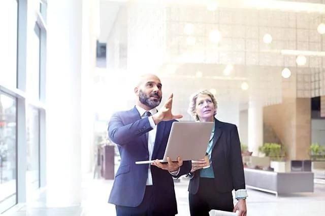 A man holding an open laptop and a woman, both in business attire, standing in a very bright room.