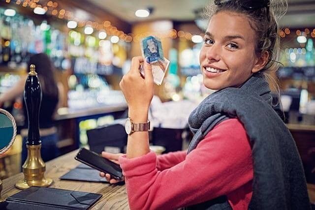 Person smiling at a bar, holding banknotes with a smartphone in hand.