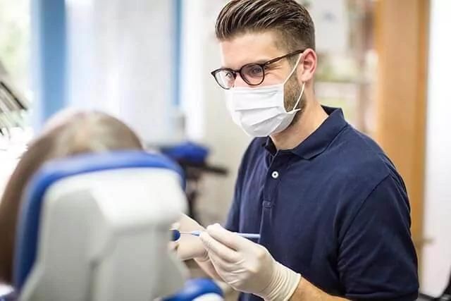 A person wearing glasses, a face mask, and gloves is sitting in a dental office, holding dental instruments. Another person, partially visible, is seated in a dental chair facing away from the camera.