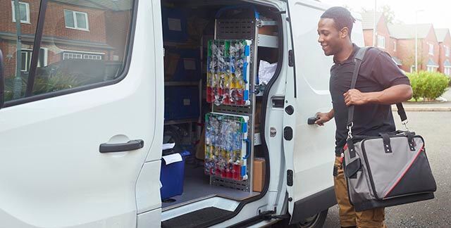 Electrician standing next to an open van holding a tool bag and visible shelves of equipment inside the van.