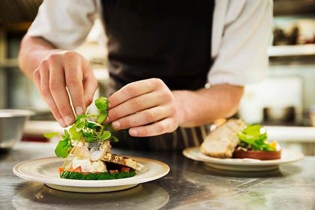Chef wearing an apron garnishing a plated dish with fresh greens in a professional kitchen.
