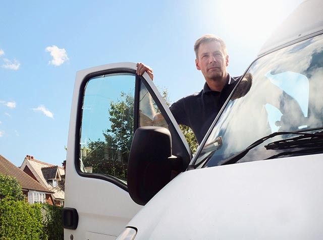 Person standing by an open van door under a sunny sky, with houses and greenery in the background.