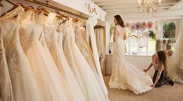 Woman in a wedding dress on a platform, with another adjusting the train in a bridal store.